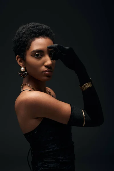 Modelo afroamericano de pelo corto y de moda en vestido, accesorios y guantes que cubren los ojos mientras posan aislados en negro, alta moda y aspecto de noche - foto de stock