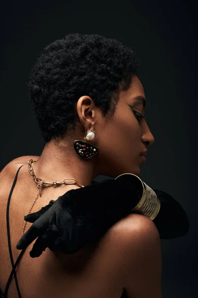 Side view of fashionable african american model in glove and modern golden chains and earring touching back and standing isolated on black, high fashion and evening look — Stock Photo