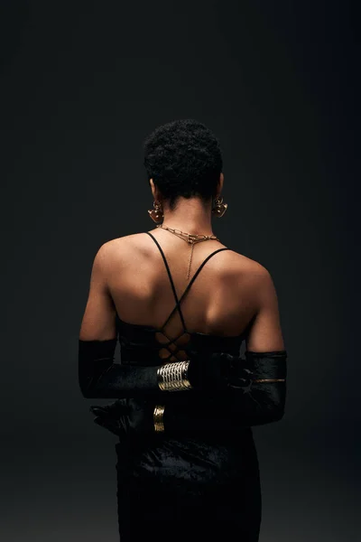 Vista posterior de la mujer afroamericana elegante y de pelo corto en guantes, vestido y accesorios modernos de pie aislado en negro, alta moda y aspecto de noche, femenino - foto de stock