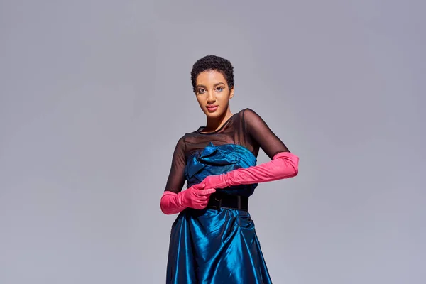 Уверенная и модная африканская американка в коктейльном платье и перчатках, смотрящая в камеру, стоя изолированно на сером, современном поколении z моды концепции — стоковое фото