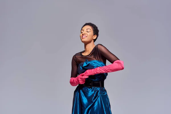Fröhliche und stilvolle junge afrikanisch-amerikanische Frau in rosa Handschuhen und Cocktailkleid, die mit geschlossenen Augen auf grauem, modernem Modekonzept der Generation z steht — Stockfoto