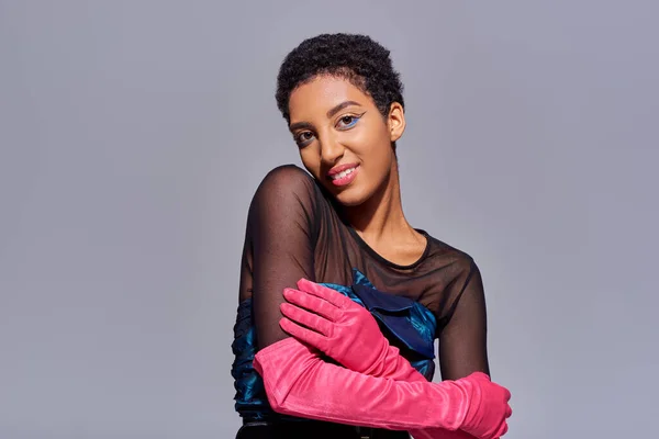 Smiling and trendy african american model in cocktail dress and pink gloves touching arm and looking at camera while posing isolated on grey, modern generation z fashion concept — Stock Photo
