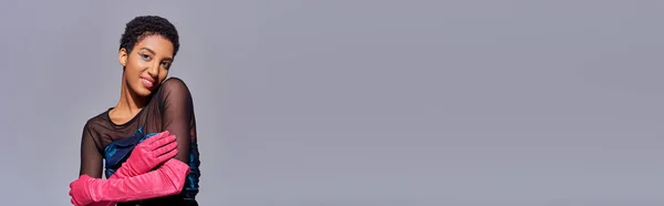 Modelo americano africano de cabelos curtos e positivos em vestido de cocktail e luvas rosa tocando braço e de pé isolado no conceito de moda z de geração moderna, cinza, banner com espaço de cópia — Fotografia de Stock