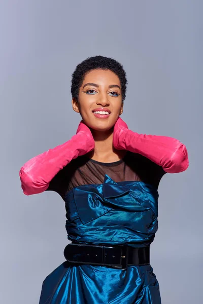 Portrait of cheerful african american woman with bold makeup posing in pink gloves and cocktail dress while touching neck and standing isolated on grey, modern generation z fashion concept — Stock Photo