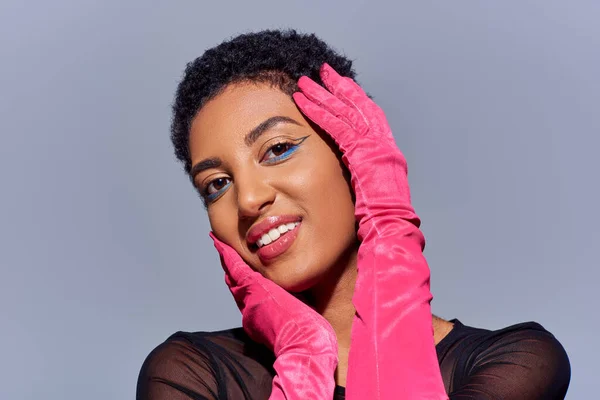 Portrait de femme afro-américaine aux cheveux courts et positifs avec un maquillage audacieux et des gants roses touchant le visage et regardant la caméra isolée sur gris, concept de mode Z génération moderne — Photo de stock