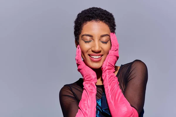 Радостная африканская американская женщина с смелым макияжем и розовыми перчатками закрывая глаза и трогательное лицо, стоя изолированы на сером, современное поколение z моды концепции — стоковое фото