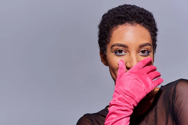 Трендовая молодая африканская женщина с смелым макияжем и розовой перчаткой, закрывающей рот и смотрящей на камеру, изолированная от серого современного поколения. — стоковое фото