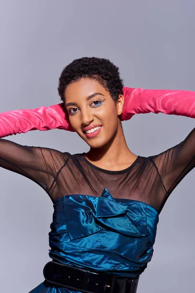 Smiling and fashionable african american woman with bold makeup wearing pink gloves and cocktail dress and touching head isolated on grey, modern generation z fashion concept — Stock Photo
