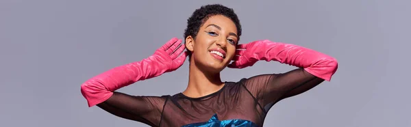 Femme afro-américaine souriante et branchée avec un maquillage audacieux portant des gants roses et posant tout en étant isolée sur le concept gris, moderne génération z, bannière — Photo de stock