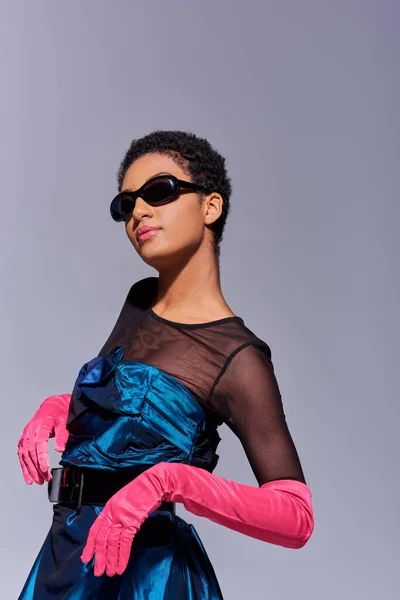 Уверенная молодая африканская американка в солнцезащитных очках, розовых перчатках и коктейльном платье позирует и стоит изолированно на сером, современном поколении z концепция моды — стоковое фото
