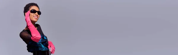 Felice donna afroamericana in guanti rosa e abito da cocktail toccare occhiali da sole e sorridente mentre in piedi isolato sul grigio, moderno concetto di moda z generazione, banner — Foto stock