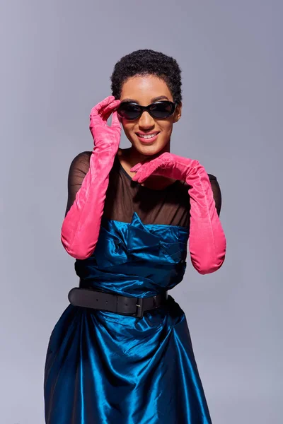 Mujer afro americana alegre y joven con el pelo corto posando en gafas de sol, guantes rosados y vestido de cóctel mientras está de pie aislado en gris, concepto de moda z generación moderna - foto de stock