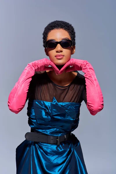 Selbstbewusste junge afrikanisch-amerikanische Frau mit Sonnenbrille, rosa Handschuhen und Cocktailkleid, die das Kinn berührt, während sie isoliert auf einem grauen Modekonzept der modernen Generation Z steht — Stockfoto