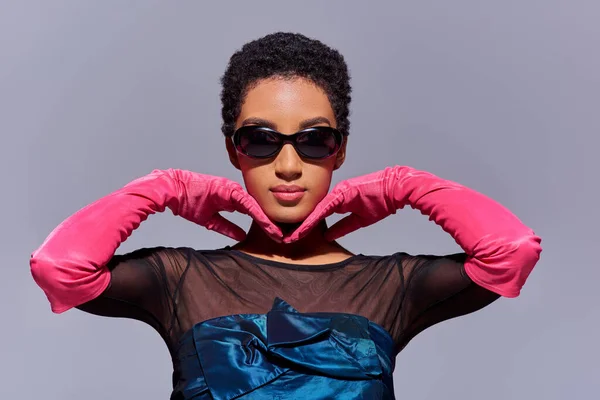 Portrait of young and short haired african american woman in sunglasses, pink gloves and cocktail dress posing isolated on grey, modern generation z fashion concept — Stock Photo