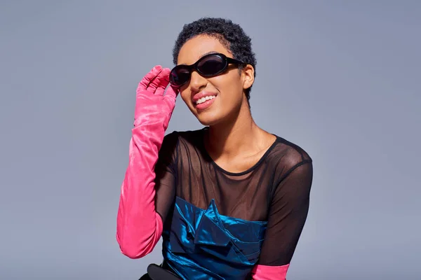 Позитивна і коротка африканська жінка, яка торкається сонцезахисних окулярів, одягаючи рожеві рукавички і вечірню сукню, будучи ізольованою на сірій концепції сучасного покоління z-моди. — стокове фото