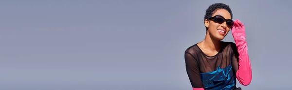 Joyeux jeune femme afro-américaine dans des lunettes de soleil élégantes, gant rose et robe de cocktail posant tout en étant isolé sur gris, concept de mode moderne de génération z, bannière — Photo de stock