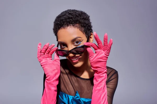 Lächelnde junge afrikanisch-amerikanische Frau mit kühnem Make-up, die Sonnenbrille berührt, während sie rosa Handschuhe trägt und sich isoliert auf grauem, modernem Modekonzept der Generation Z kleidet — Stockfoto