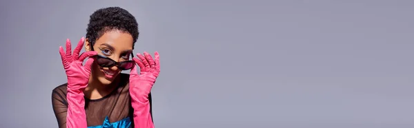 Mulher americana africana alegre com maquiagem ousada e cabelo curto posando em óculos de sol e luvas rosa isolado em cinza, conceito de moda z geração moderna, banner com espaço de cópia — Fotografia de Stock