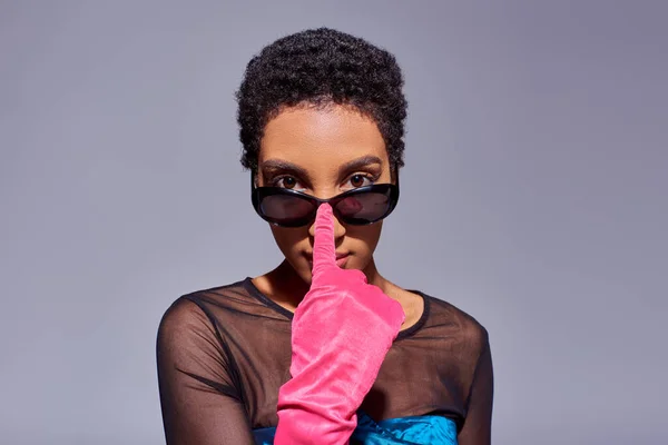 Retrato de mujer afroamericana de pelo corto en guante rosa tocando gafas de sol y mirando a la cámara mientras está de pie aislado en gris, concepto de moda z generación moderna - foto de stock