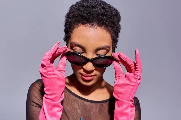 Jeune et confiante femme afro-américaine touchant des lunettes de soleil et portant des gants roses tout en posant et debout isolé sur gris, concept de mode de génération z moderne — Photo de stock