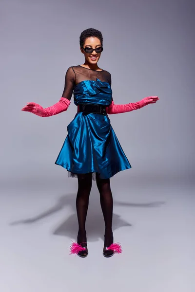 Pleine longueur de femme afro-américaine gaie en lunettes de soleil, robe de cocktail et gants roses posant dans des chaussures à plumes tout en se tenant debout sur fond gris, concept de mode moderne génération z — Photo de stock