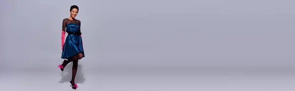 Modello afroamericano sorridente in abito da cocktail, guanti rosa e tacchi con piume che guardano la fotocamera mentre si trova su sfondo grigio, concetto di moda z di generazione moderna, banner — Foto stock