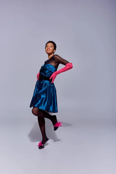 Mujer afroamericana de moda en vestido de cóctel, guantes de color rosa y tacones con plumas tocando las caderas y sonriendo mientras está de pie sobre fondo gris, concepto de moda z generación moderna - foto de stock