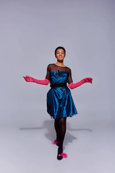 Longitud completa de alegre modelo afroamericano en guantes de color rosa, vestido de cóctel y tacones con plumas caminando y mirando a la cámara sobre fondo gris, concepto de moda z generación moderna - foto de stock