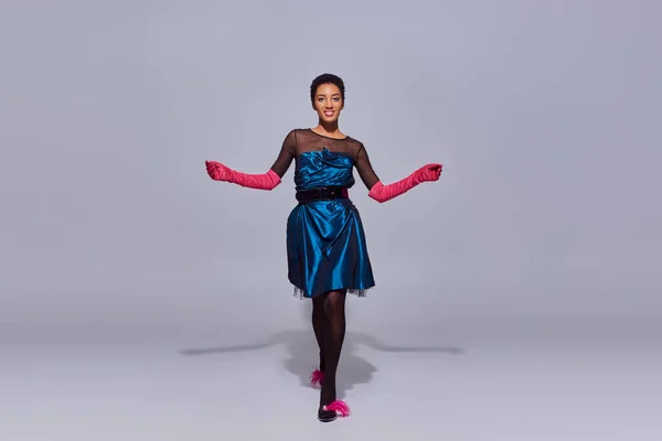 Mujer afroamericana sonriente y de pelo corto en vestido de cóctel, guantes rosas y tacones con plumas caminando sobre fondo gris, concepto de moda z generación moderna - foto de stock