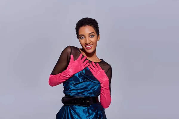 Портрет позитивної і стильної афроамериканської моделі зі сміливим макіяжем в рожевих рукавичках і коктейльній сукні, що дивиться на камеру ізольовано на сірому, сучасному поколінні концепції моди Z — стокове фото