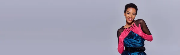 Retrato del modelo afroamericano sonriente en guantes rosados y vestido de cóctel sonriendo y mirando a la cámara aislada en gris, concepto de moda z generación moderna, bandera - foto de stock
