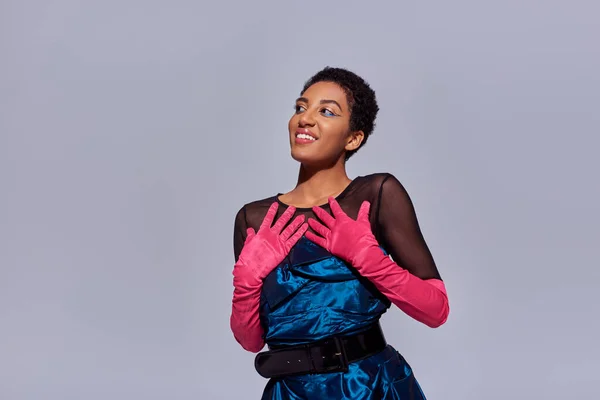 Positive african american model with short hair posing in pink gloves and cocktail dress while standing with hands near chest isolated on grey, modern generation z fashion concept — Stock Photo