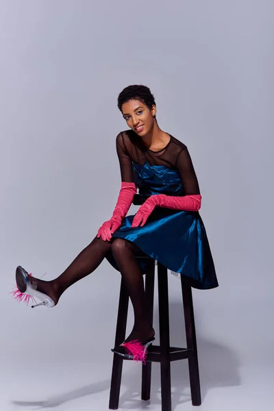 Lächelndes afrikanisch-amerikanisches Model in rosa Handschuhen, Cocktailkleid und High Heels mit Federn auf Stuhl auf grauem Hintergrund, Modekonzept der modernen Generation z — Stockfoto