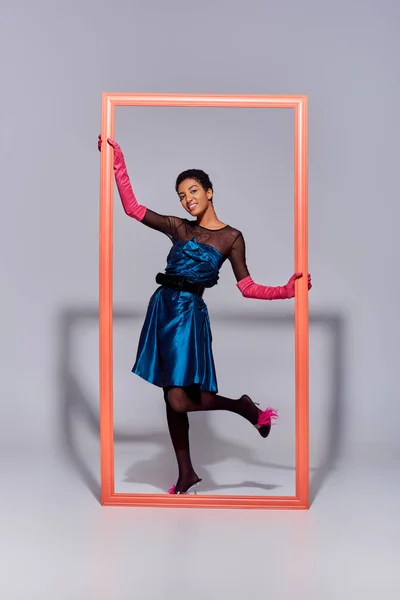 Полная длина улыбающейся и стильной африканской модели в коктейльном платье и каблуках, глядя на камеру рядом с кадром и стоя на сером фоне, современное поколение z моды концепции — стоковое фото