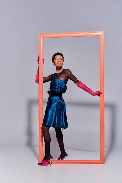 Trendy and smiling young african american woman in pink gloves, cocktail dress and feathered shoes standing near frame and posing on grey background, modern generation z fashion concept — Stock Photo