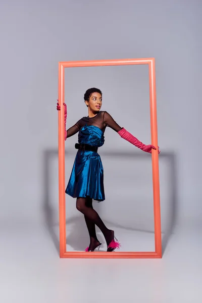 Трендовая юная африканская модель в коктейльном платье, розовых перчатках и туфлях на шпильках позирует и стоит рядом с рамкой на сером фоне, концепция моды современного поколения Z — стоковое фото