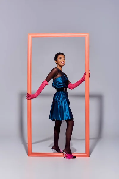 Joyful african american model in evening dress, pink gloves and heels with feathers looking at camera near frame on grey background, modern generation z fashion concept — Stock Photo