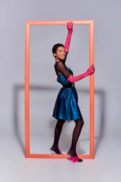 Happy and short haired african american woman in pink gloves, cocktail dress and heels with feathers touching frame on grey background, modern generation z fashion concept — Stock Photo