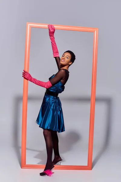 Full length of trendy african american woman in pink gloves, feathered shoes and cocktail dress smiling at camera while standing near frame on grey background, modern generation z fashion concept — Stock Photo