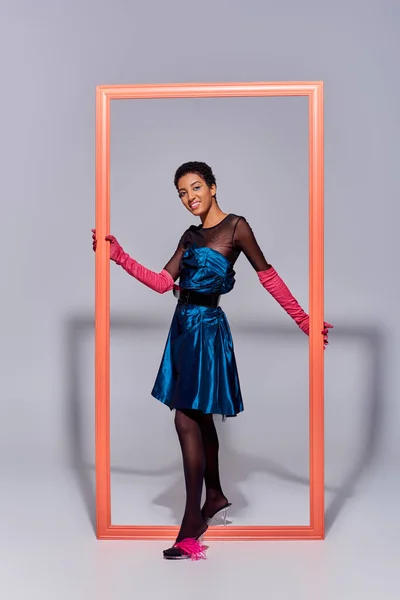 Весела афроамериканська модель в рожевих рукавичках, підбори з пір'ям і коктейльна сукня, що стоїть біля рамки і позує на сірому фоні, концепція моди сучасного покоління z — стокове фото