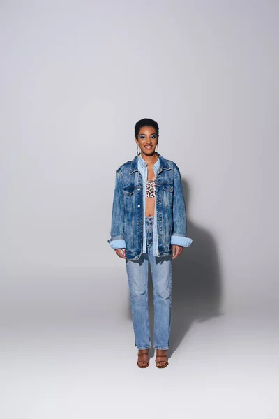 Full length of trendy and short haired african american model in denim clothes and jeans smiling at camera while posing and standing on grey background, denim fashion concept — Stock Photo
