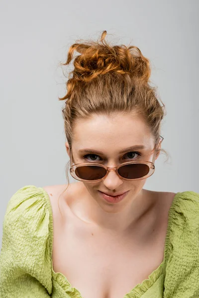 Jeune femme rousse tendance et moderne en chemisier vert et lunettes de soleil regardant la caméra tout en étant isolé sur fond gris, concept de protection solaire tendance, modèle de mode — Photo de stock