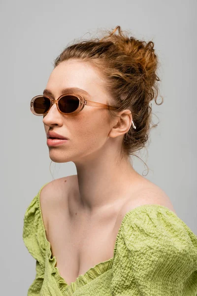 Портрет стильної молодої рудої волохатої жінки в зеленій блузці і сучасних сонцезахисних окулярах, що позують, стоячи ізольовано на сірому фоні, концепція модного захисту від сонця — стокове фото