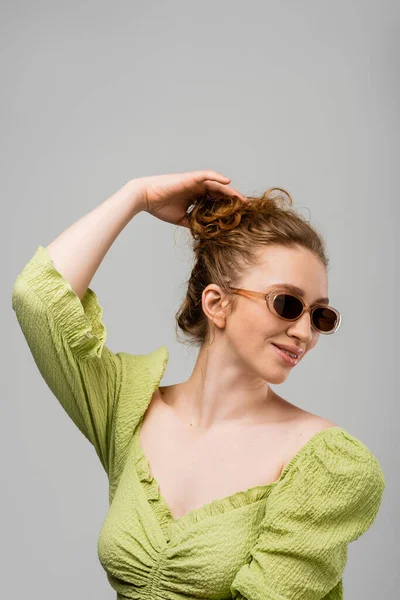 Sorridente giovane donna rossa in estate camicetta verde e occhiali da sole toccare i capelli mentre in piedi isolato su sfondo grigio, concetto di protezione solare alla moda, modello di moda — Foto stock