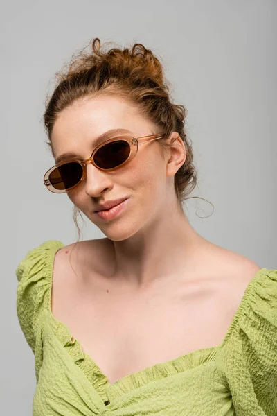 Portrait of young redhead and freckled woman in green blouse and stylish sunglasses standing and posing isolated on grey background, trendy sun protection concept, fashion model — Stock Photo