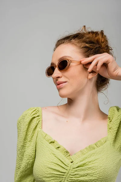Mujer pelirroja sonriente y moderna en blusa verde tocando gafas de sol elegantes mientras está de pie aislado sobre fondo gris, concepto de protección solar de moda, modelo de moda - foto de stock
