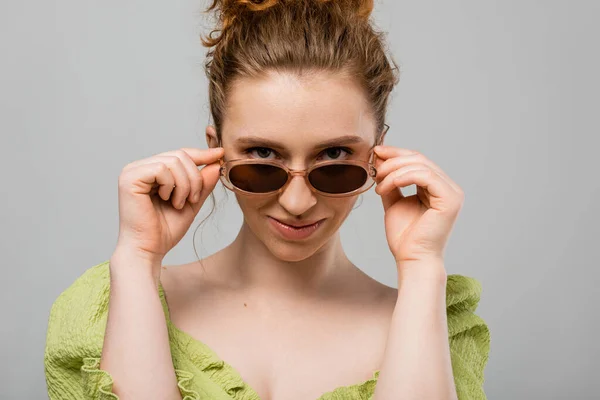 Stylish young redhead woman with natural makeup wearing green blouse and touching sunglasses while looking at camera isolated on grey background, trendy sun protection concept, fashion model — Stock Photo