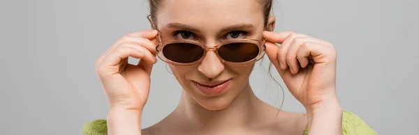 Портрет сучасної молодої жінки з натуральним макіяжем, що торкається стильних сонцезахисних окулярів і дивиться на камеру ізольовано на сірому фоні, концепція модного захисту від сонця, банер, модель моди — стокове фото