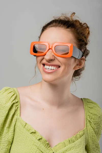 Porträt einer fröhlichen und stilvollen rothaarigen und sommersprossigen Frau mit Sonnenbrille und grüner Bluse, die die Augen schließt und isoliert auf grauem Hintergrund steht, trendiges Sonnenschutzkonzept, Modemodel — Stockfoto