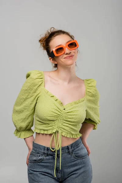Mujer joven sonriente en blusa verde, jeans y gafas de sol posando y mirando hacia otro lado mientras está de pie aislada sobre un fondo gris, modelo de moda, concepto de protección solar de moda - foto de stock